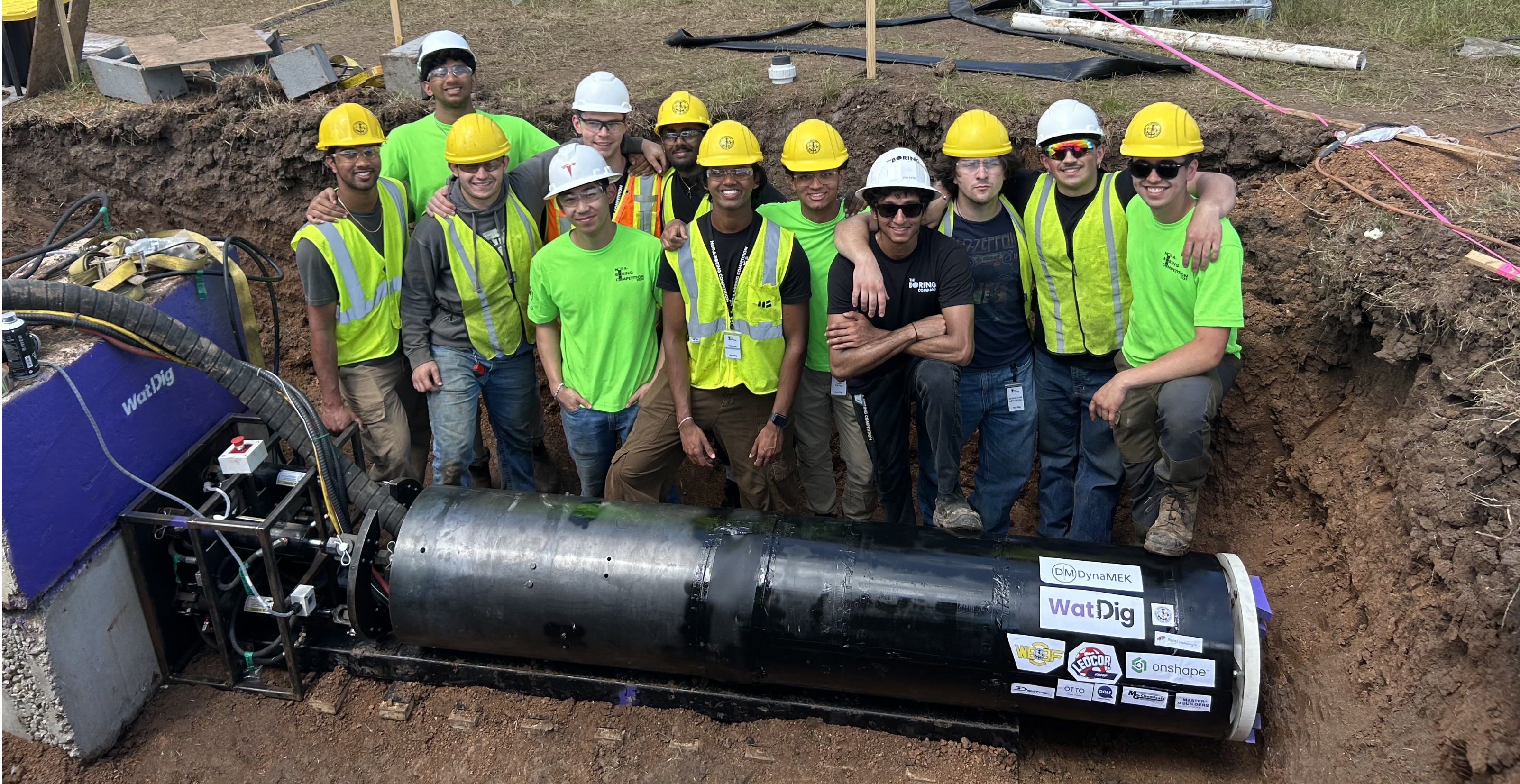 The WatDIG team in hard hats and hi-vis vests standing in an excavated launch pit behind their black micro tunnel boring machine, with the WatDIG logo on the machine and on the jacking frame beside it.